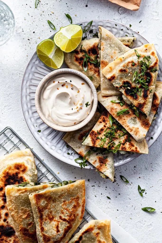 A view looking down on a white plate filled with pieces of stuffed flatbread served with whipped yogurt, sitting beside a wire cooling rack with fresh cooked bolani. 