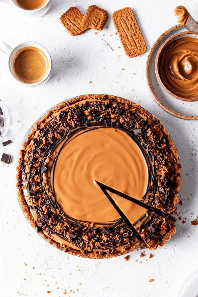 A chocolate cookie tart with one clean slice cut, beside a cup of espresso, a small bowl of cookie butter and some speculoos cookies. 