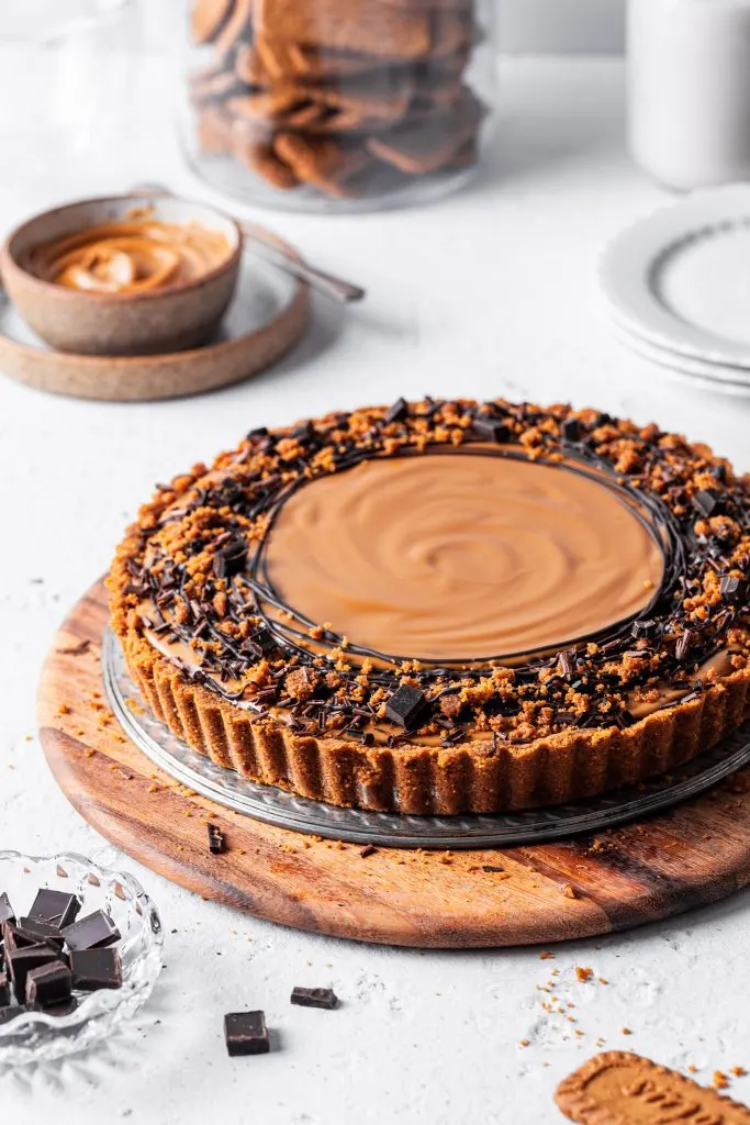 Fully assembled chocolate biscoff tart is garnished with drizzles of dark chocolate, with a jar of Biscoff cookies and a small bowl of cookie butter in the background. 