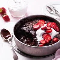 A round cake pan filled with a partially served chocolate cake exposing chocolate sauce on the bottom. Garnished with whipped cream and sliced strawberries.