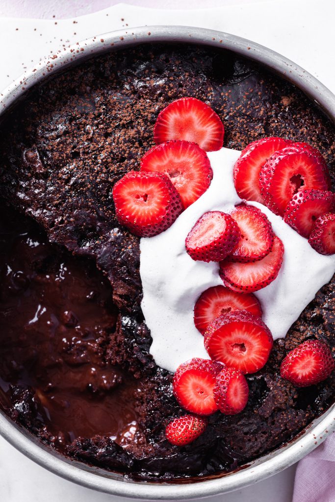 Close up of the self-saucing chocolate strawberry pudding cake with a serving missing so that both the cake and the sauce are visible. The top of the vegan dessert is topped with non-dairy whip and fresh sliced strawberries. 