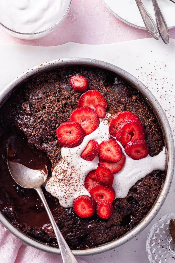 View looking down on a baking dish filled with chocolate cake baked on top of sauce. One scoop has been removed, and the top is garnished with vegan cream and sliced strawberries. 