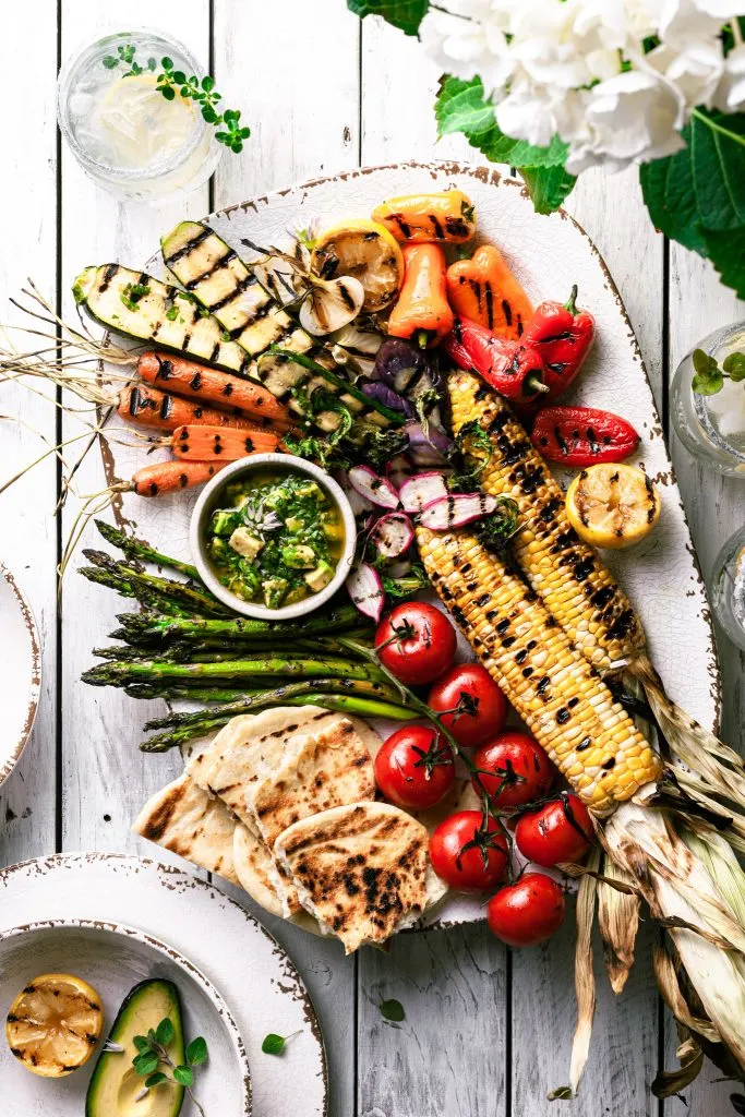 A white platter filled with grilled vegetables and a small bowl of chimichurri sauce on a white picnic table.