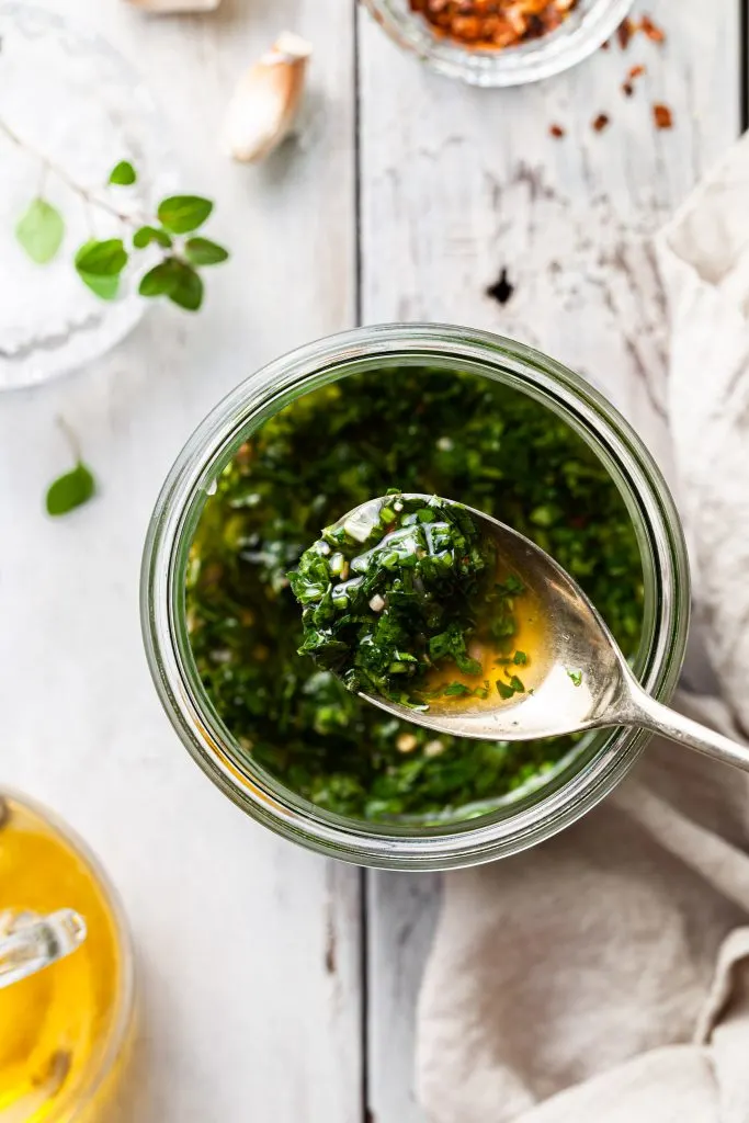 A picture with a shallow depth of filed focusing on chimichurri on a spoon hovering over a jar filled with the green sauce. 