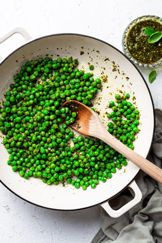 A white enamel skillet filled with sautéed peas covered in basil pesto. 