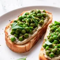 Two slices of toast on a white plate, topped with whipped vegan ricotta and green peas covered in pesto. A lemon wedge is in the background beside a glass of water.