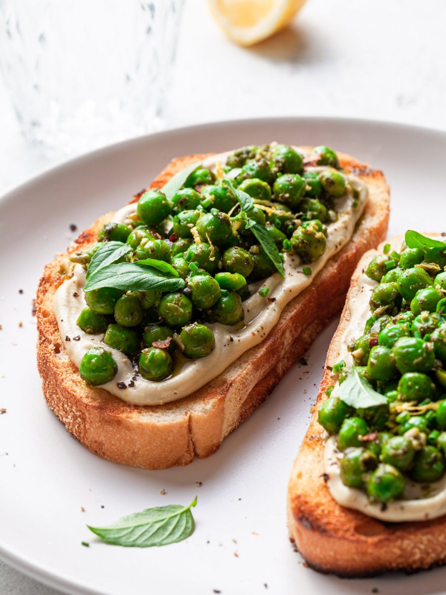 Two slices of toast on a white plate, topped with whipped vegan ricotta and green peas covered in pesto. A lemon wedge is in the background beside a glass of water.