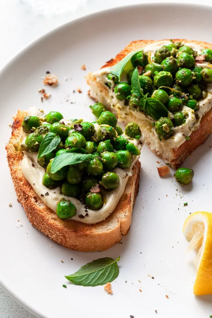 A slice of toast cut in half, covered in a creamy spread and topped with green peas covered in a basil sauce.