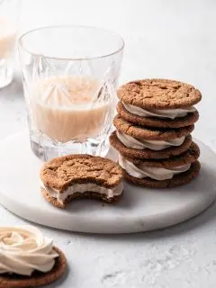 A stack of molasses sandwich cookies beside a glass of vegan eggnog all on a white marble plate