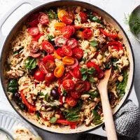 A white cast iron skillet filled with orzo, chickpeas, tomatoes and other Greek inspired herbs and veggies.