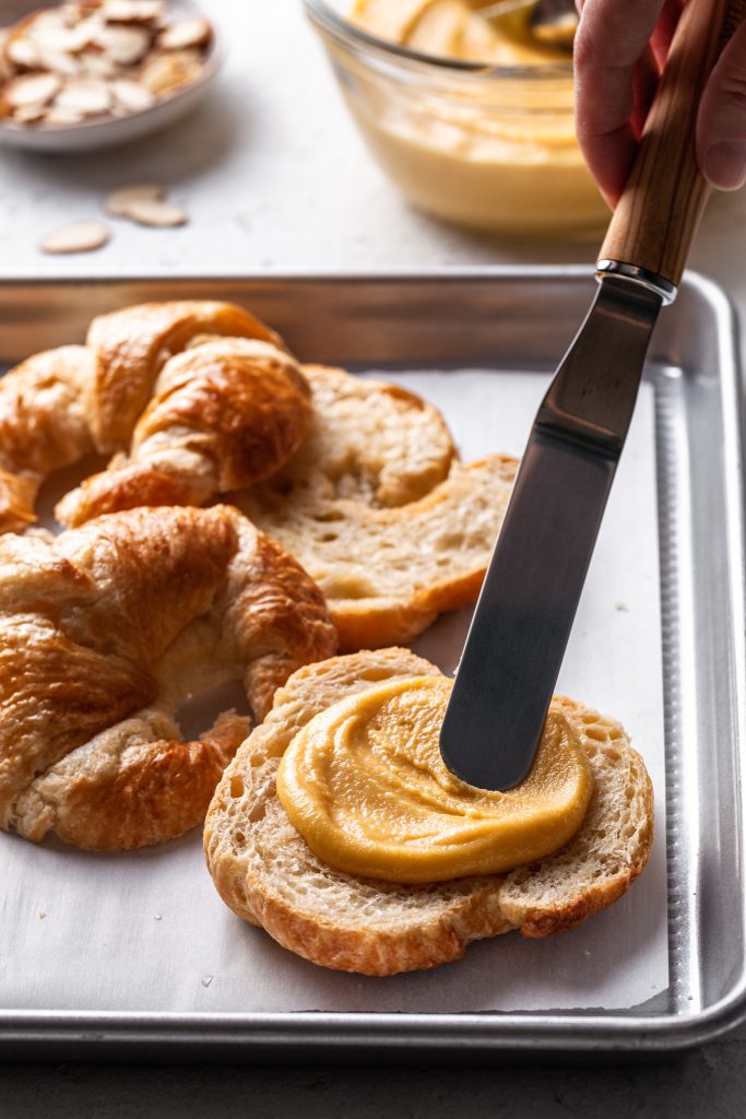 An offset spatula spreading frangipane onto the bottom cut side of a croissant. 