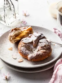 Two almond croissants on a cream plate, one is broken open to reveal the creamy filling.