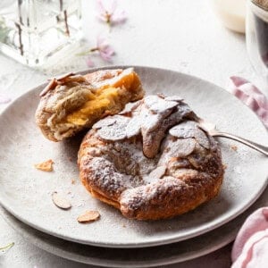Two almond croissants on a cream plate, one is broken open to reveal the creamy filling.