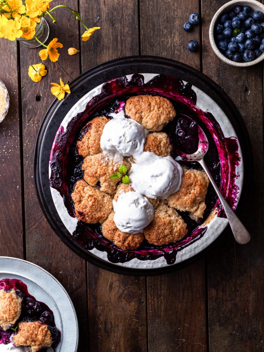 Baked blueberry cobbler in a white round baking dish, with 3 scoops of vanilla ice cream served on top.