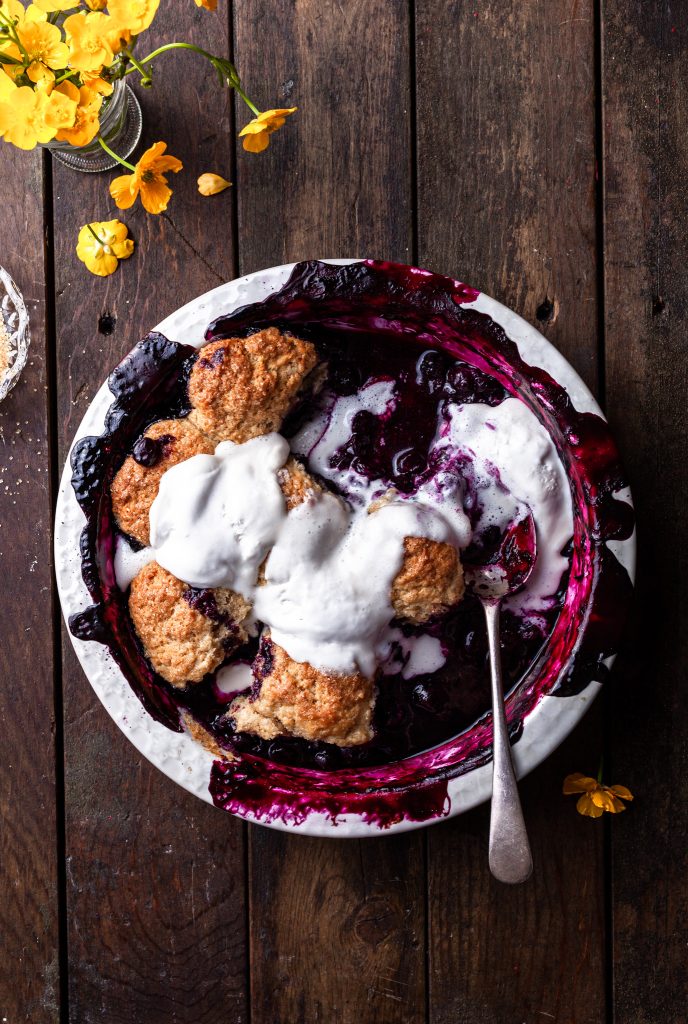 Blueberry cobbler in a white baking dish with a few servings missing, topped with melting vanilla ice cream.