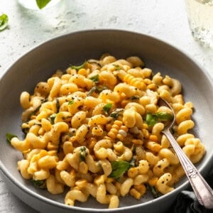 A light grey coup plate filled with cavatappi pasta coated in a creamy sauce studded with corn kernels and basil.
