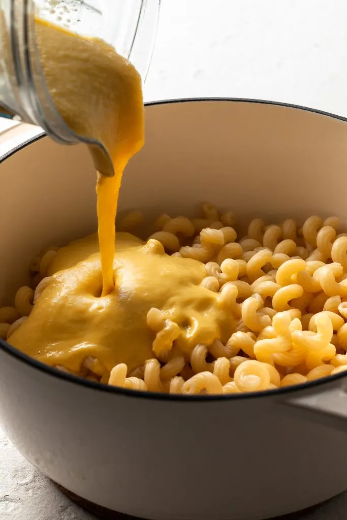 A creamy yellow sauce pouring out of a blender jug into a pot filled with cooked pasta noodles.