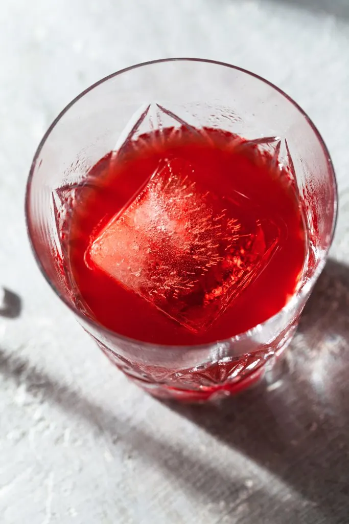 Angled view into a highball glass filled with a red drink and a square ice cube. 