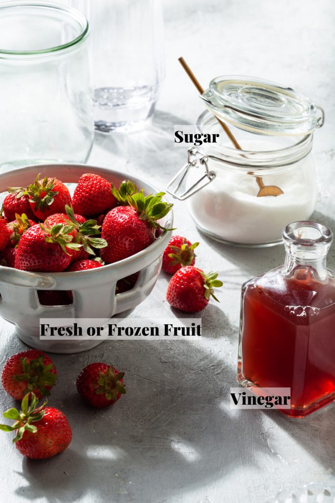 A grey counter lined with a colander of fresh berries, a glass canister of sugar and a bottle of vinegar.
