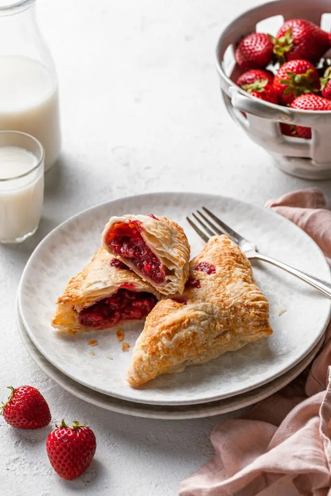 Two turnovers on a speckled cream plate, one is whole and the other is cut in half to display the red berry filling inside. A basket of strawberries is in the background.