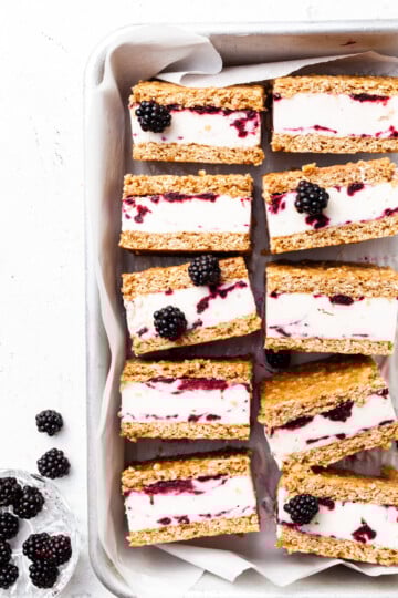 Baking tin lined with white parchment, filled with ice cream sandwiches that contain and purple swirl through the white ice cream layer.