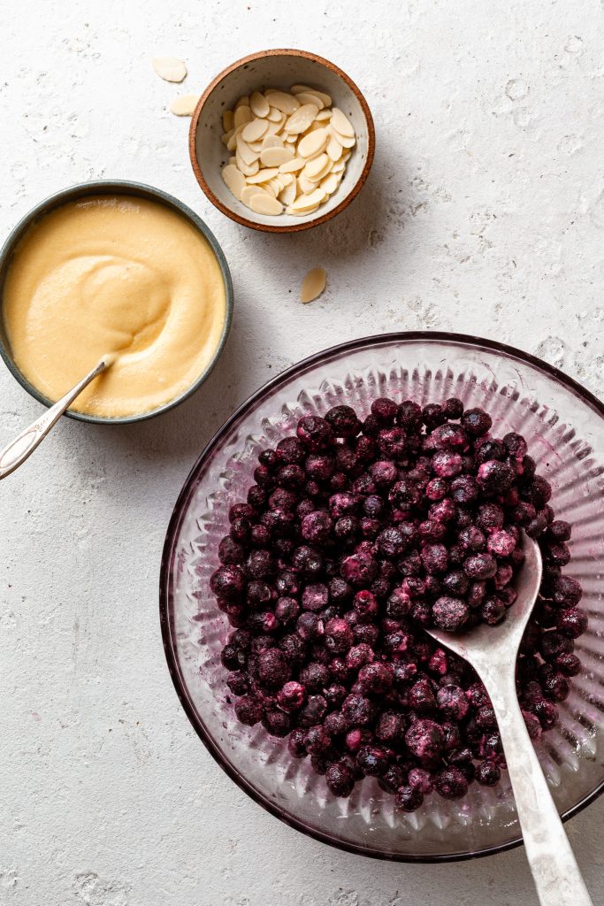 A bowl of berries, a bowl of yellow creamy mixture, and a small bowl of slivered almonds on a white countertop.
