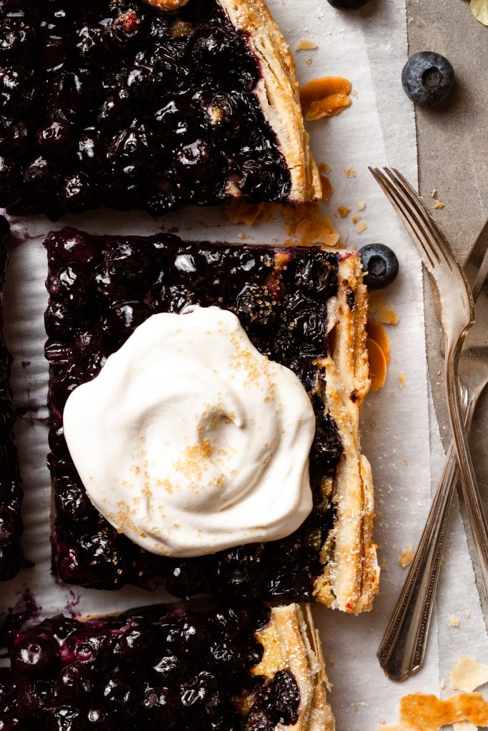 Large slice of blueberry pie topped with whip cream and sprinkled with coarse sugar on a piece of parchment paper.
