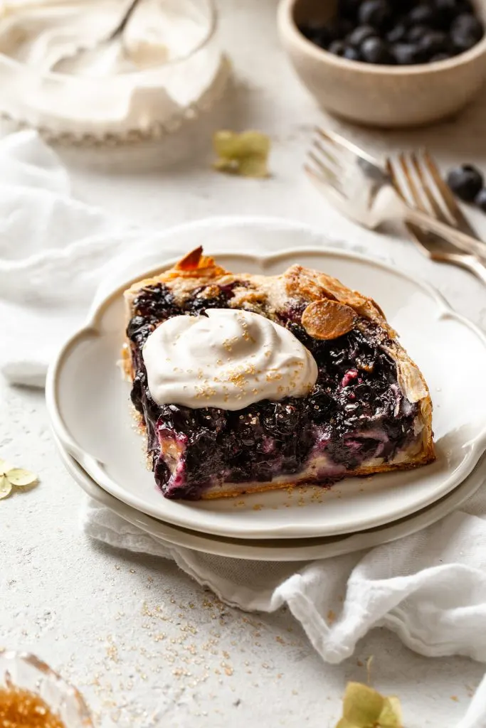 Slice of blueberry pie on a white plate, topped with shipped cream.