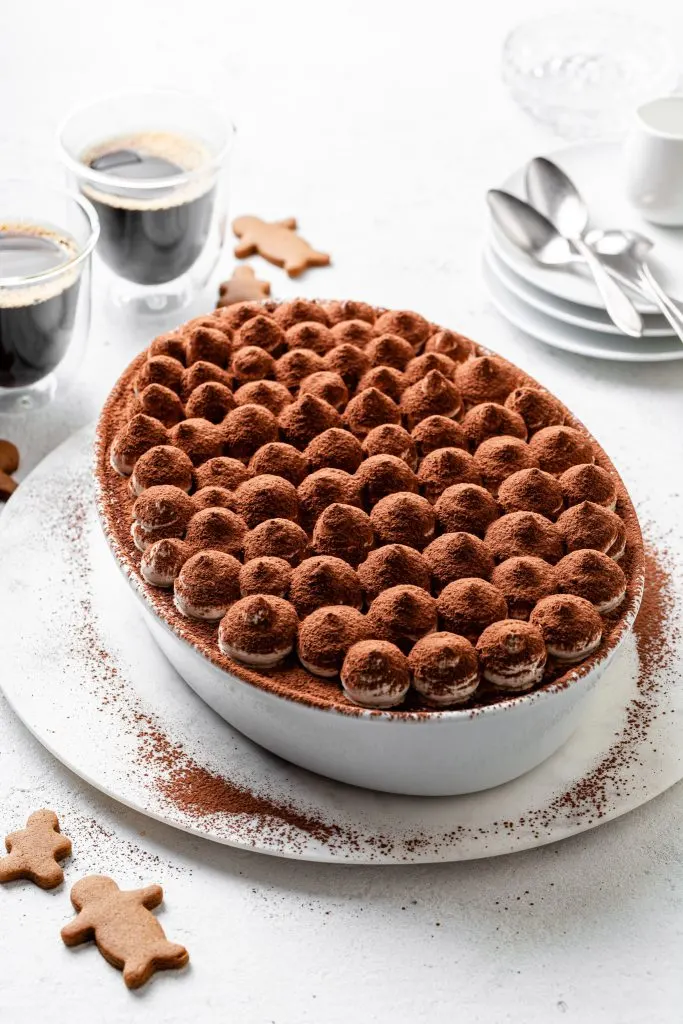 A white oval dish filled with tiramisu on a white counter, with glass cups of coffee beside gingerbread people cookies in the background.