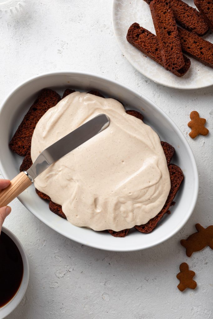 A creamy filling being spread over a layer of gingerbread cookies in a white baking dish. 
