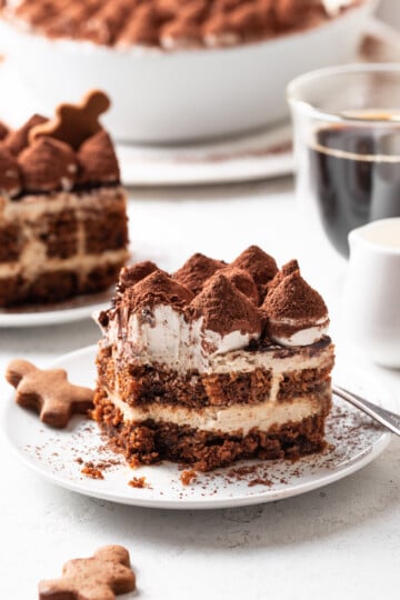 A serving of tiramisu missing a bite on a white plate beside a gingerbread cookie.