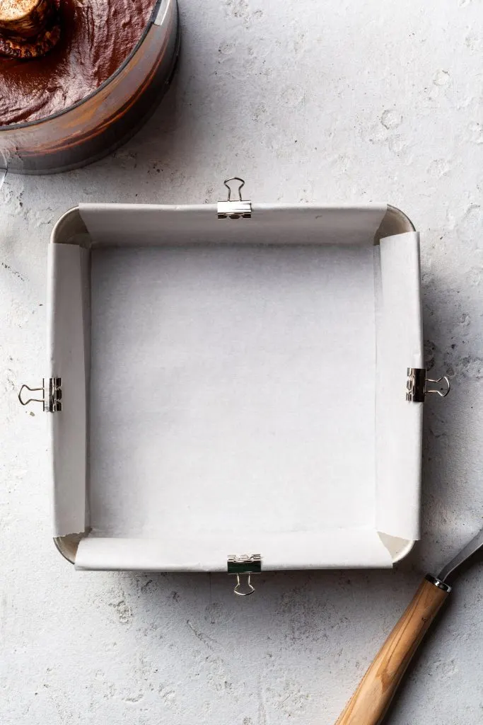 A square baking pan lined with white parchment paper secured with small silver metal clips.