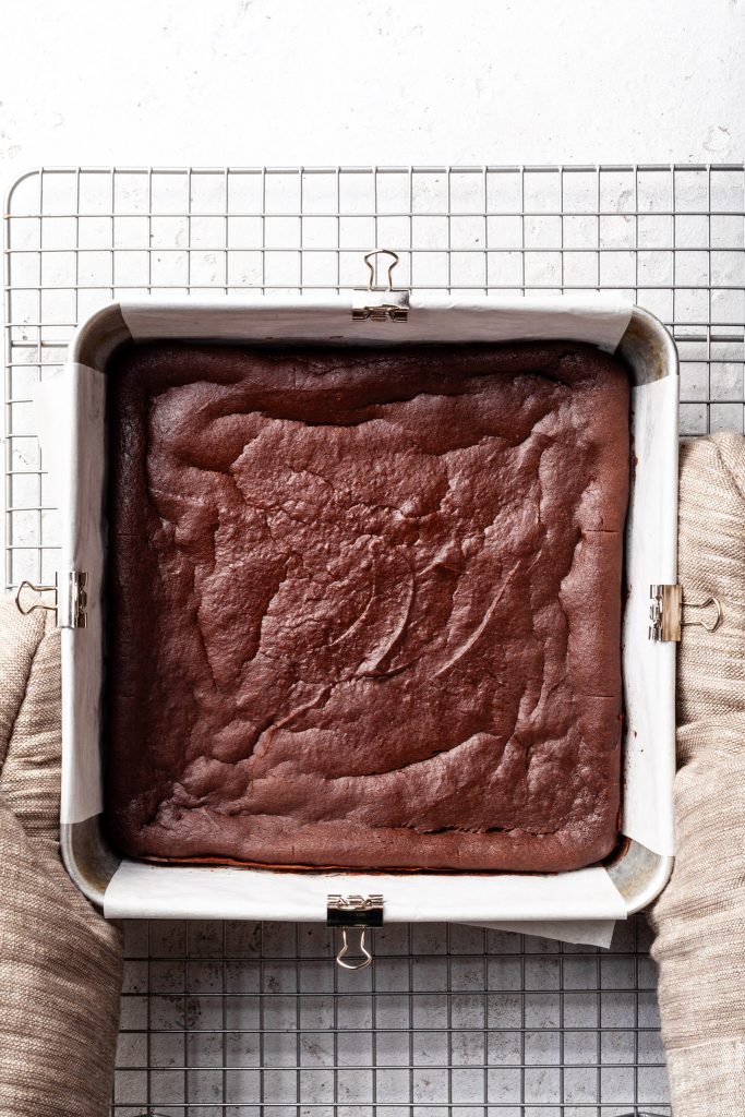 Hands with oven mitts on, holding a square tin of chocolate brownies over a cooling rack. 