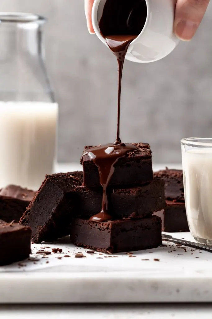 A square marble platter displays a batch of sliced brownies stacked along side a bottle of milk. A small white jug pours chocolate sauce over 3 of the brownies. 